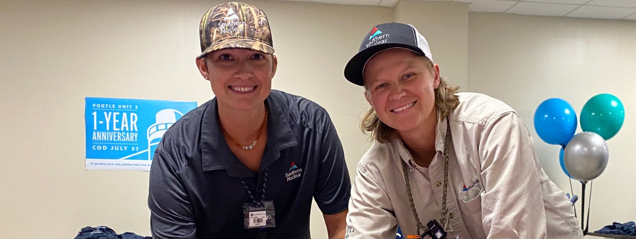 Two Southern Nuclear employees smile at a Vogtle Unit 3 celebration event, wearing company hats and standing near balloons and a display sign.