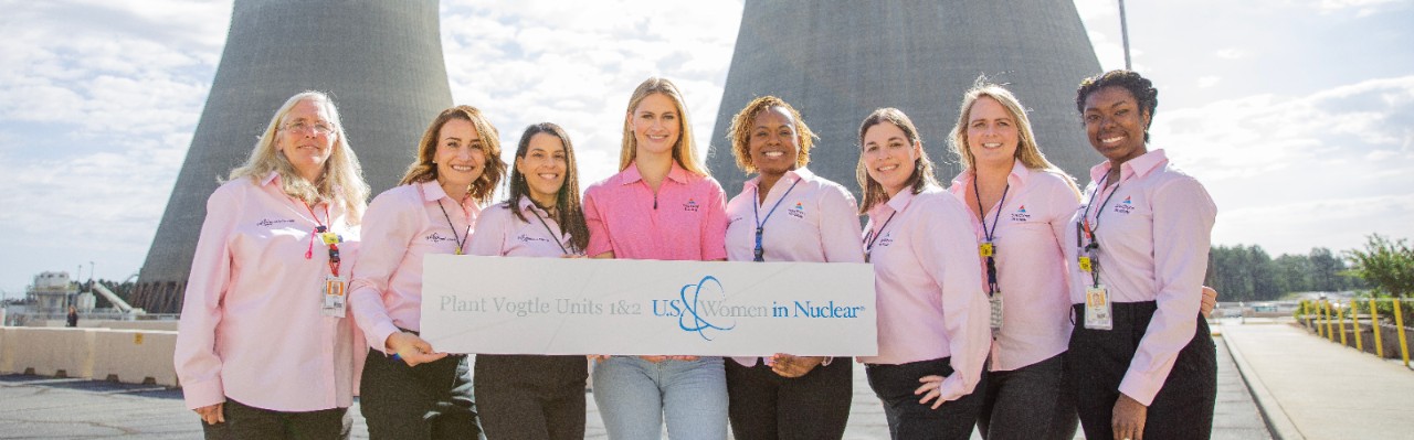Group of women from Southern Nuclear’s Plant Vogtle team pose proudly outside cooling towers, holding a sign for U.S. Women in Nuclear.