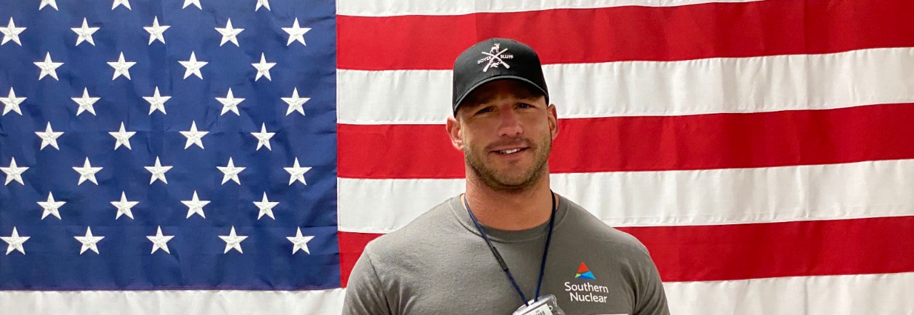 Southern Nuclear employee in branded shirt and hat stands proudly before an American flag, symbolizing nuclear power plant jobs and service.