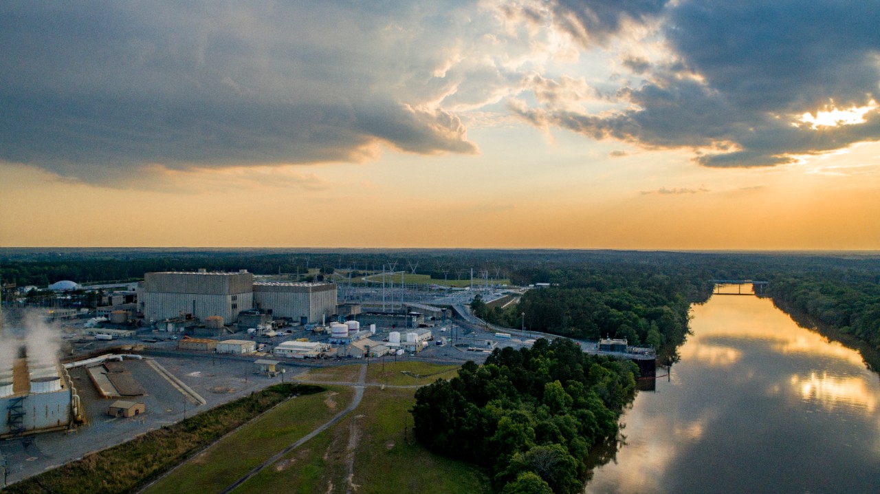 Southern Nuclear's Plant Hatch facility beside a calm river at sunset, with industrial buildings, power lines, and sky casting warm reflections.