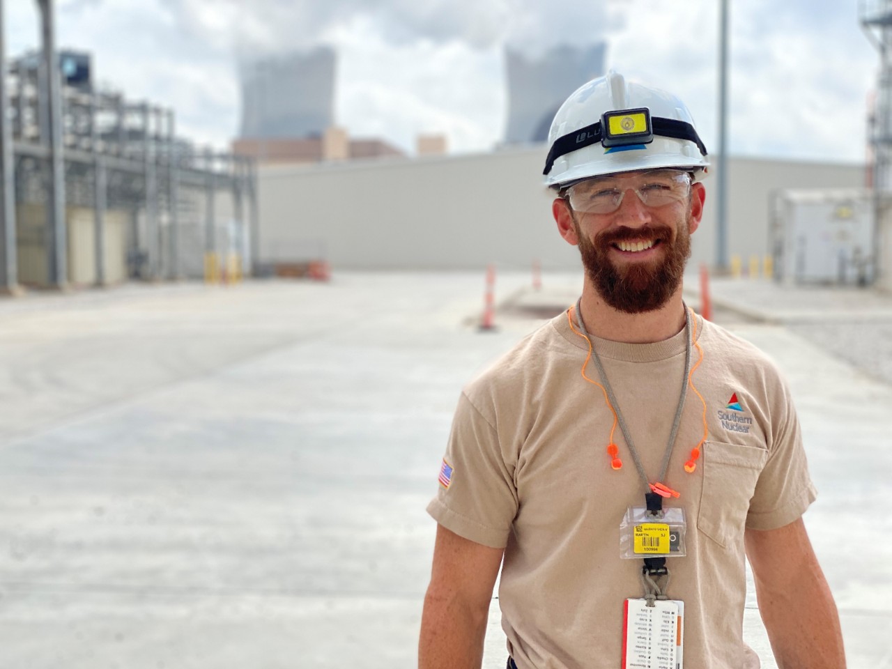 A smiling Plant Vogtle worker in safety gear stands outdoors near equipment and buildings, with the power plant’s cooling towers in the background.