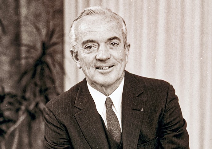 A formal portrait of Alvin W. Vogtle in a dark suit and tie, seated and smiling in front of a curtain with leafy plants in the background.
